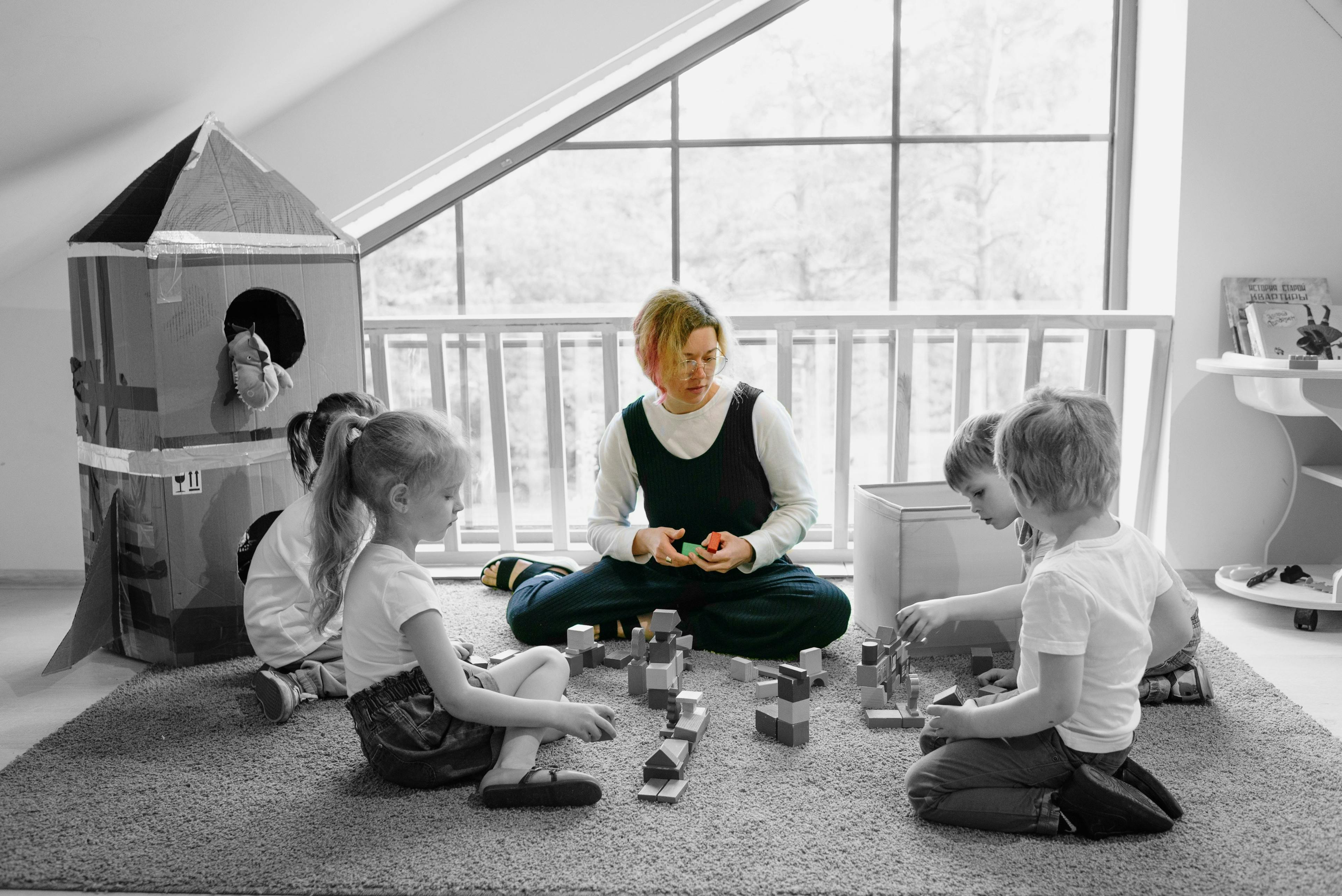 Female nursery teacher sits on rug playing blocks with four children sitting around her
