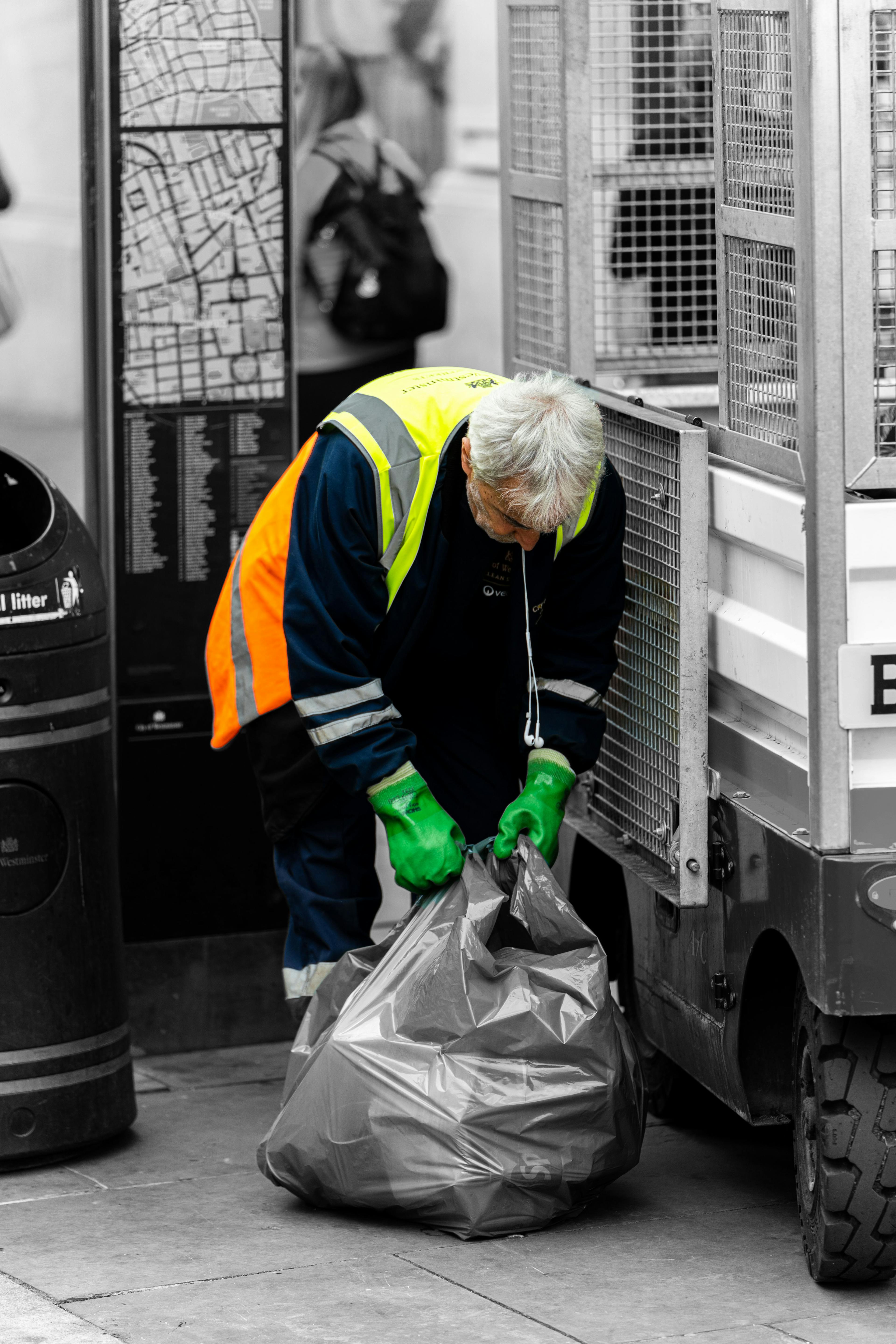 Refuse worker in protective clothing and gloves looks down as he ties up a rubbish bag