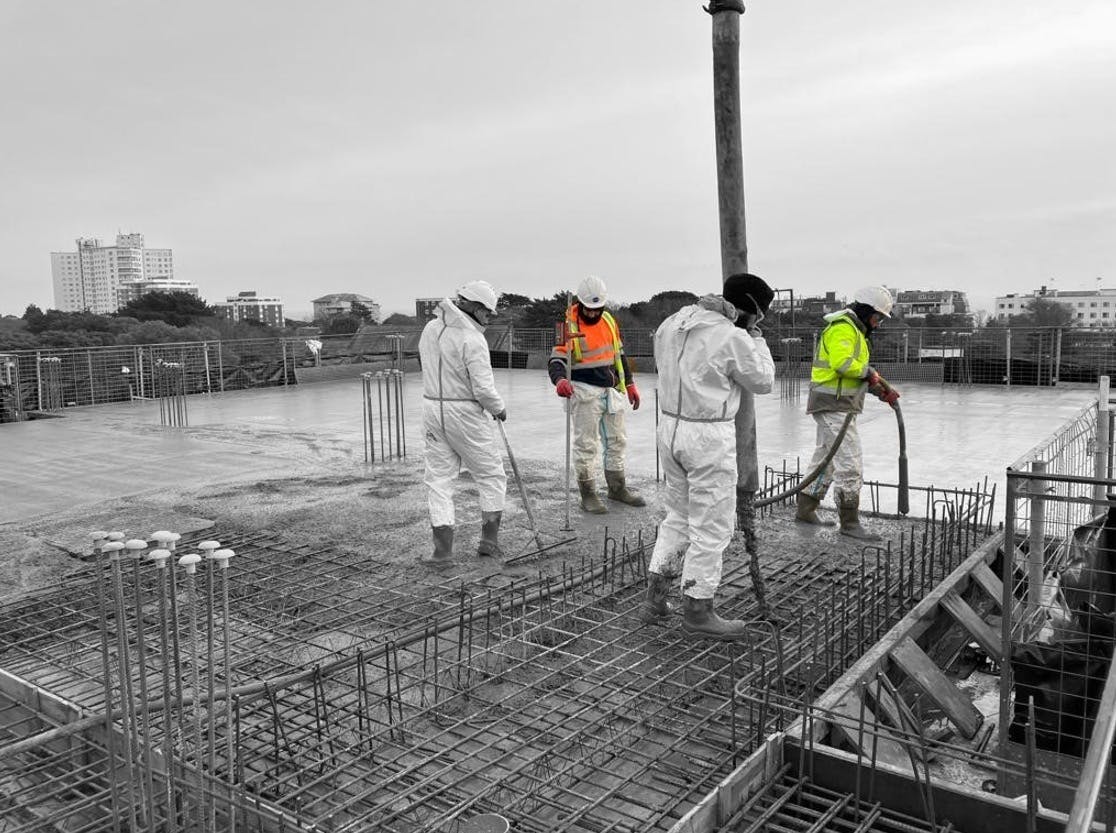 Four construction workers wearing full protective clothing pouring cement onto the top of a new building