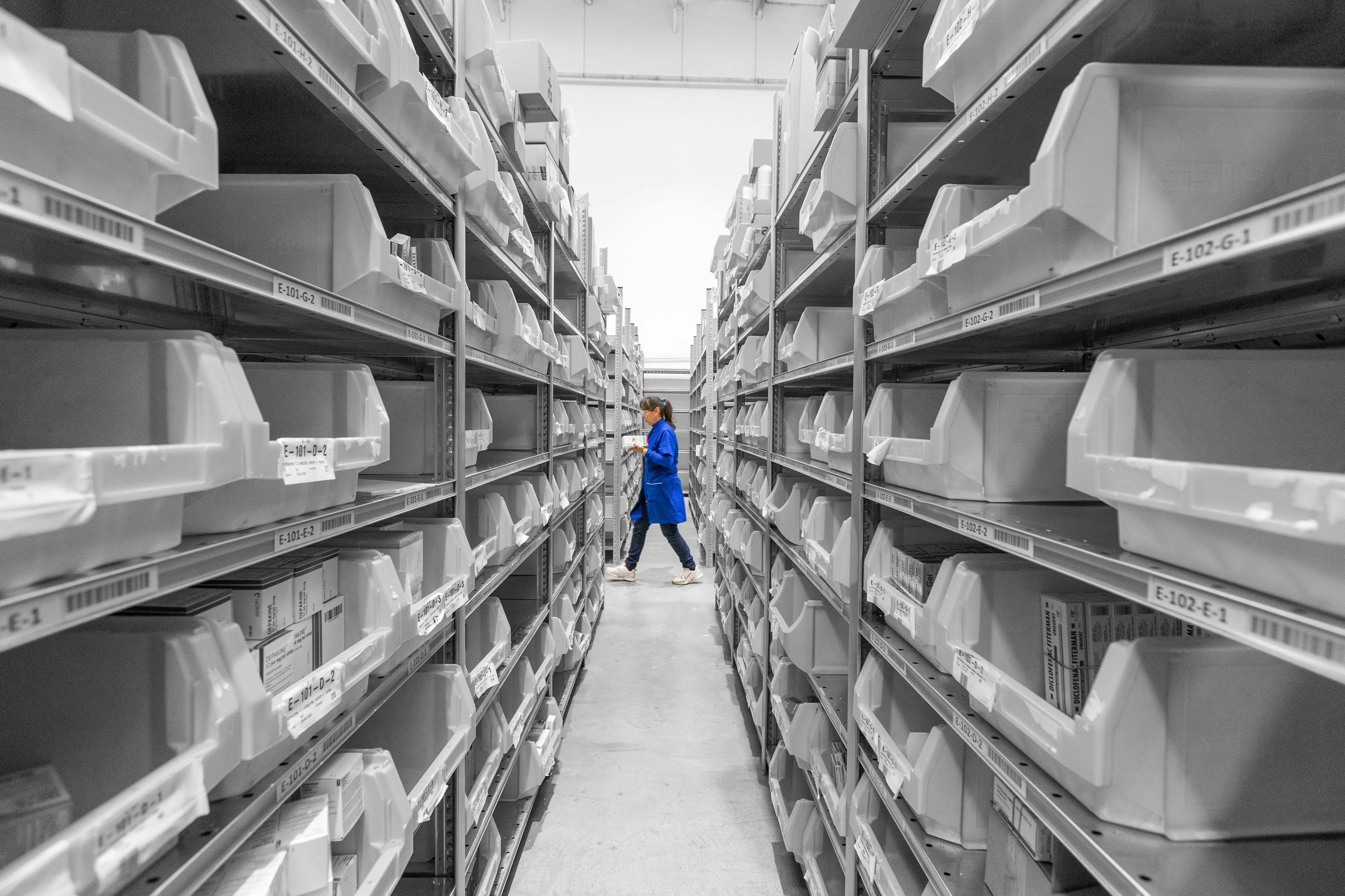 Female warehouse worker wearing blue jacket walking along row of empty shelves