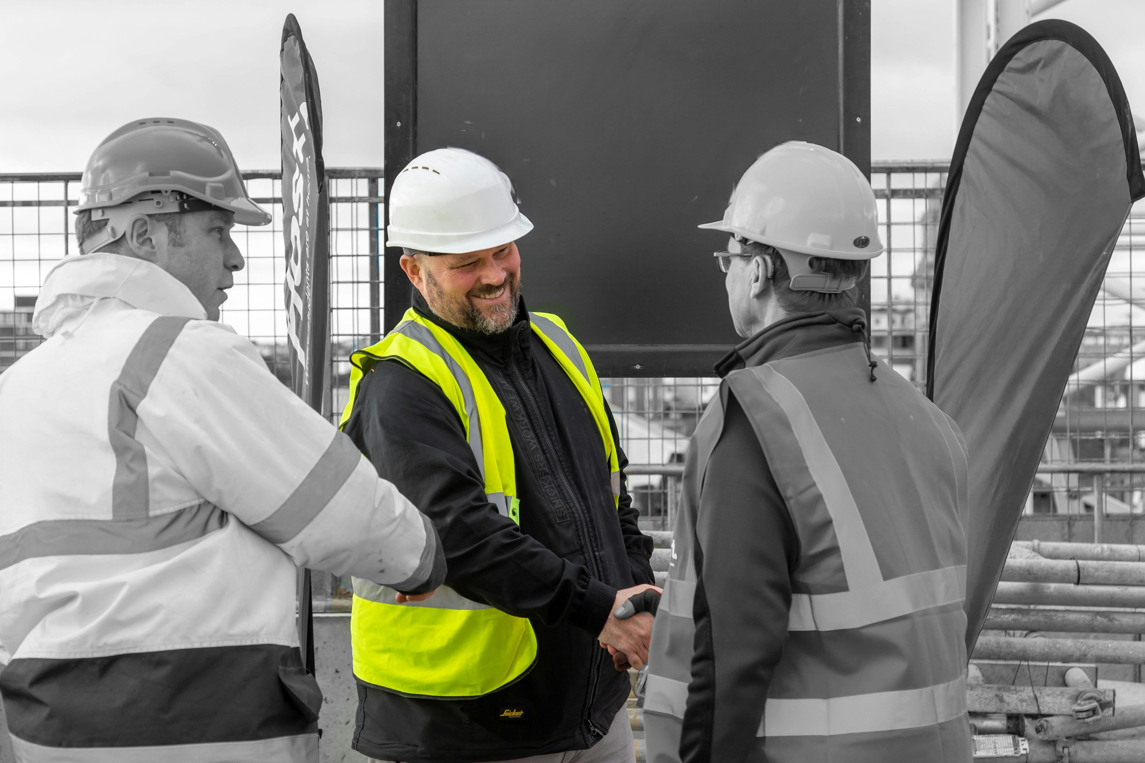 Two construction workers wearing hard hats shaking hands whilst another worker looks on