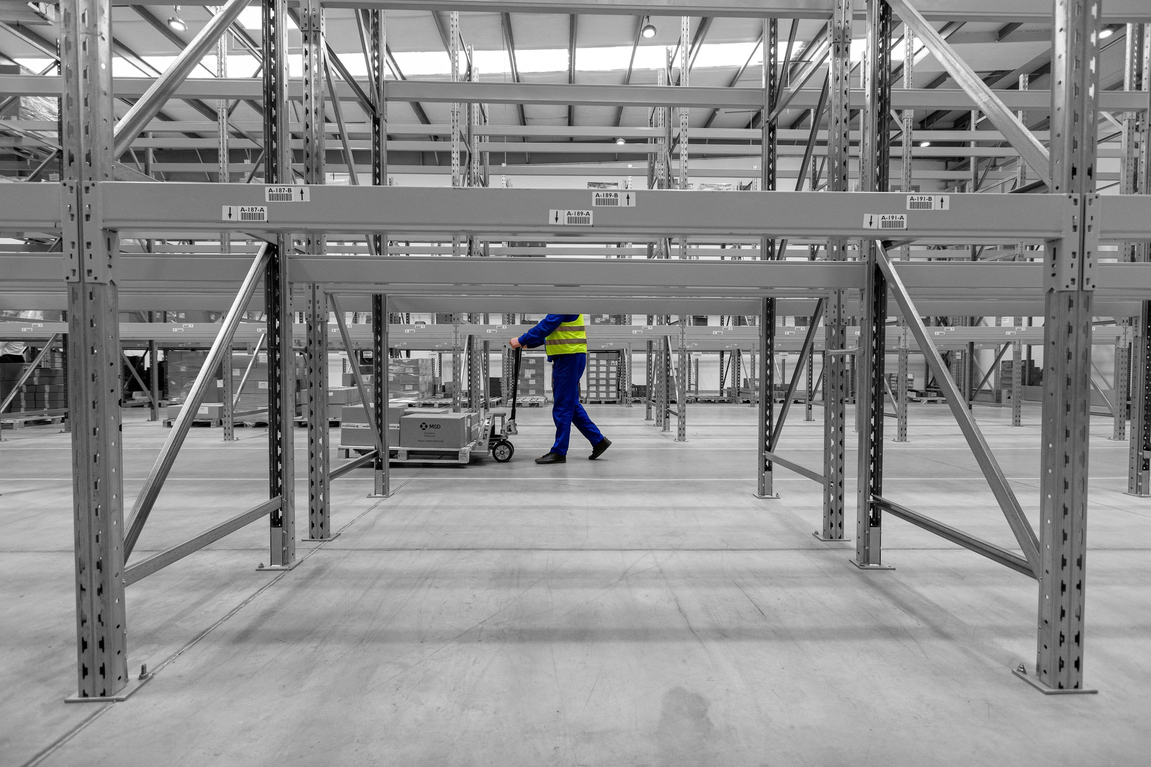Logistics worker in hi vis jacket pushing warehouse trolley along warehouse aisle
