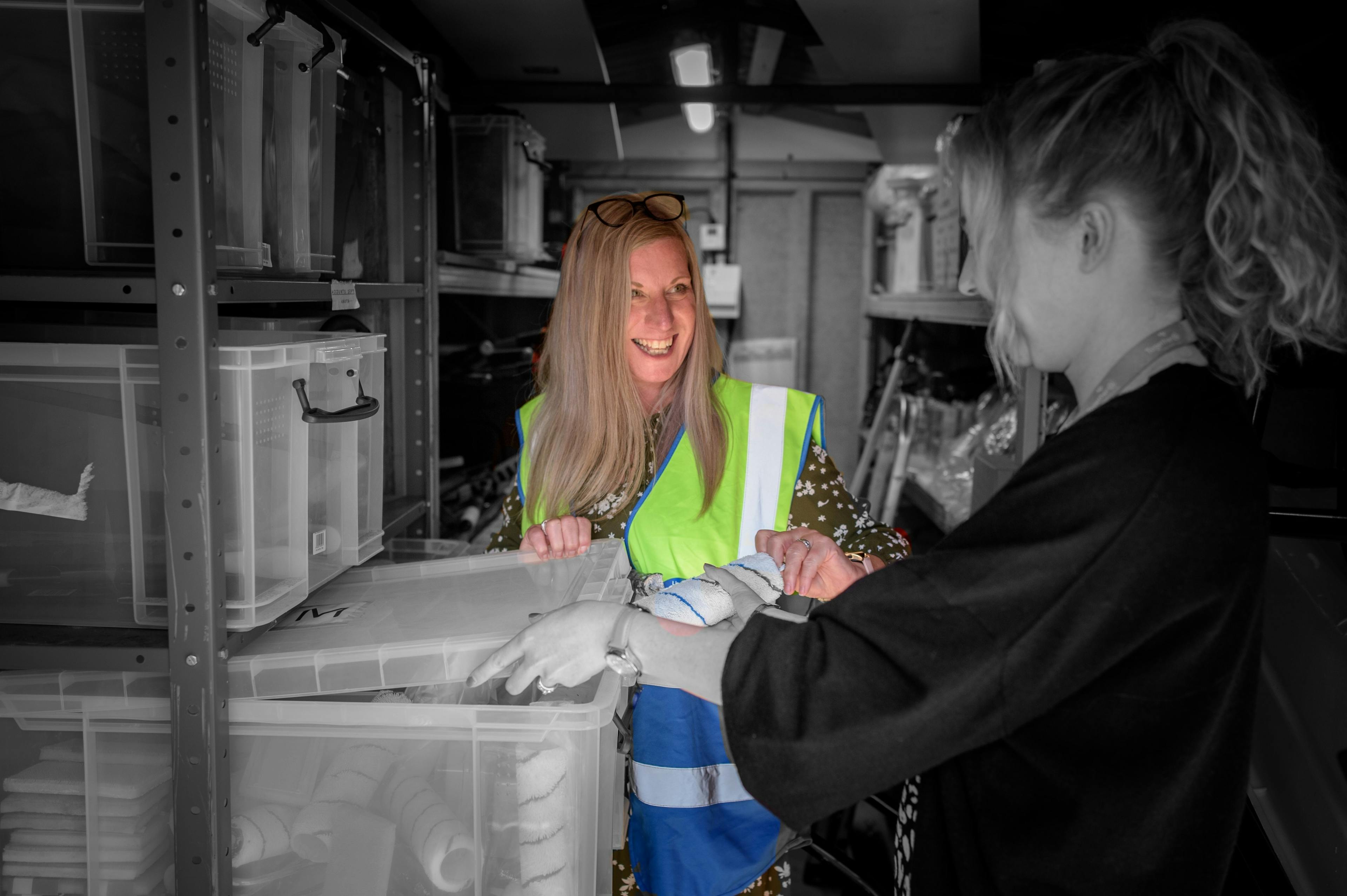 Two women talking in a store room, one wearing a hi vis jacket and smiling 