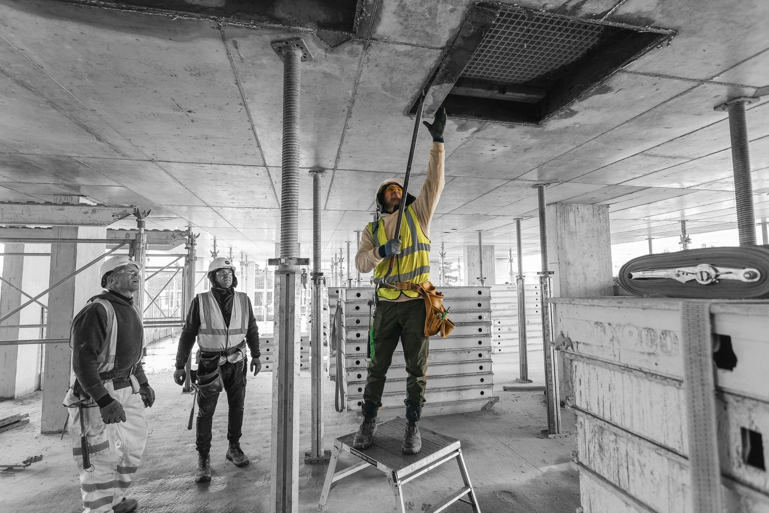 Two male construction workers on partially complete construction site watching a third worker remove a piece of wood from a concrete ceiling using a crowbar