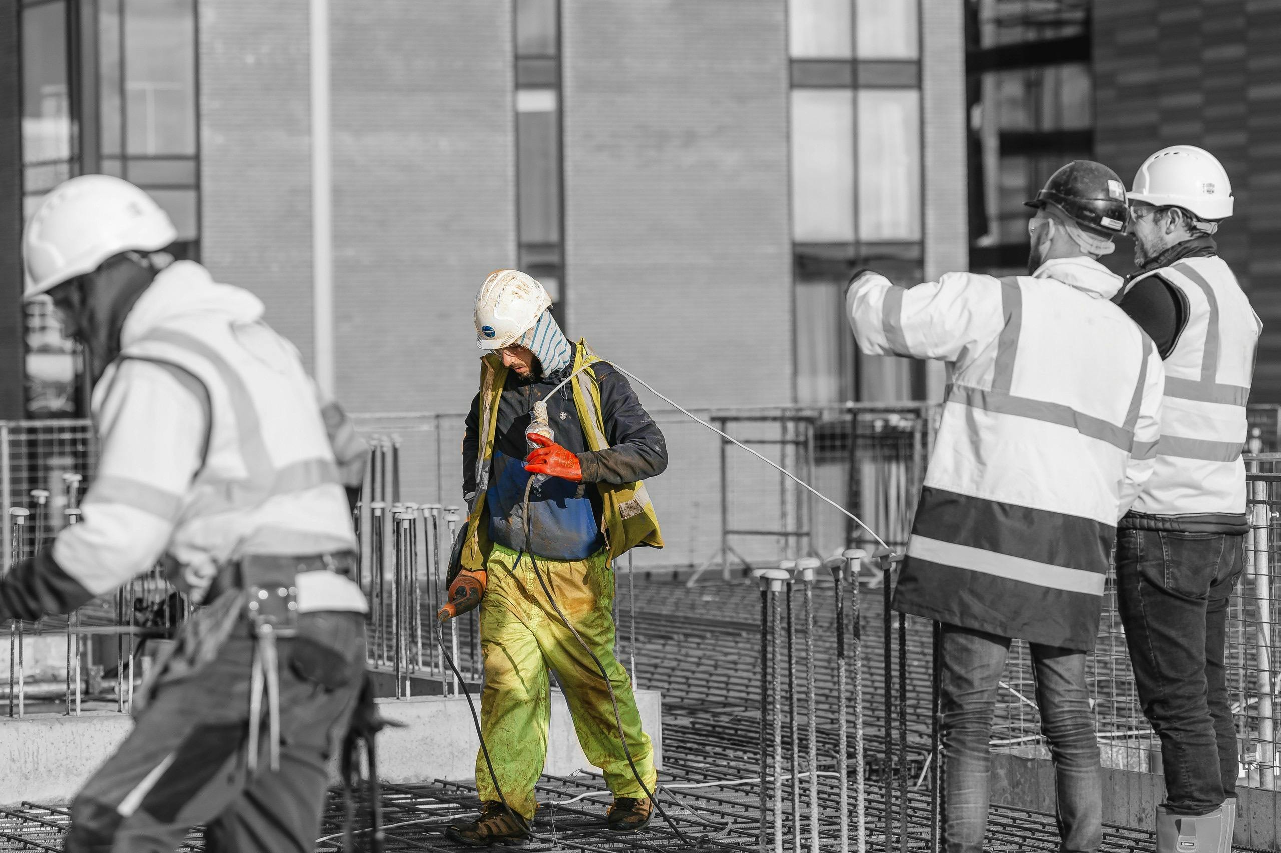 Two workers on a building site being watched by two men talking in protective clothing