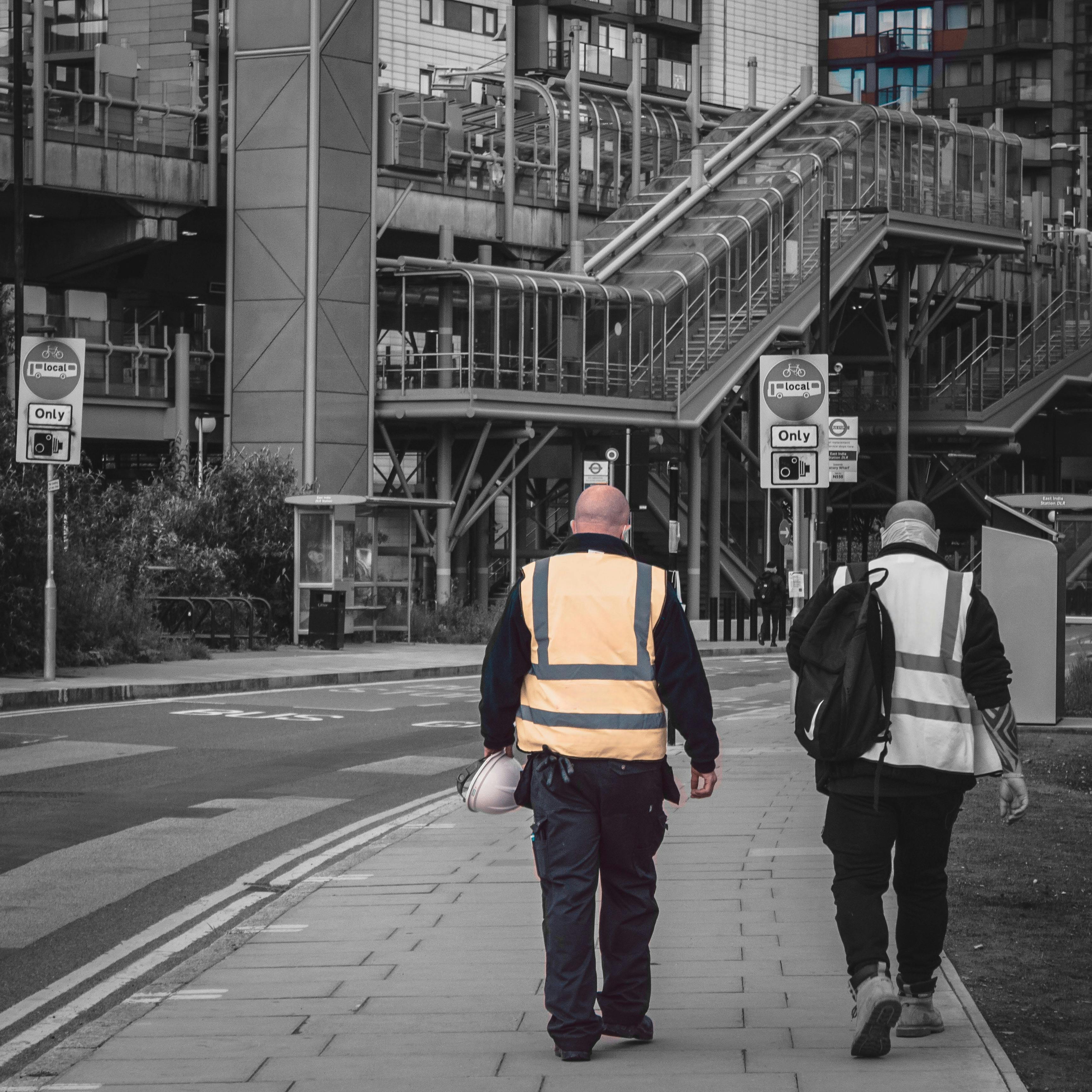 Two workers in hi vis jackets walk away from camera towards train station in London