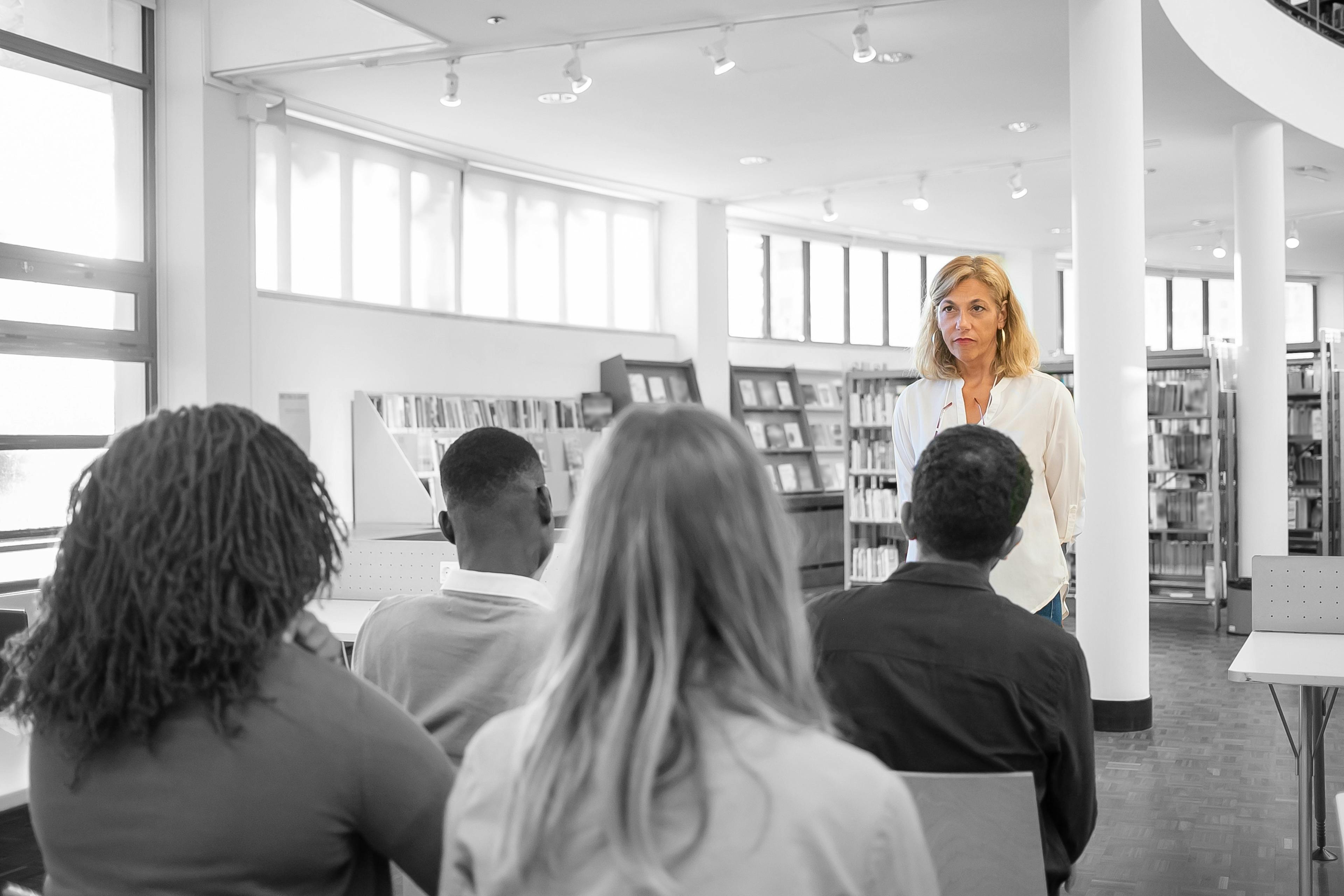 Female teacher stands in front of seated students in a library 