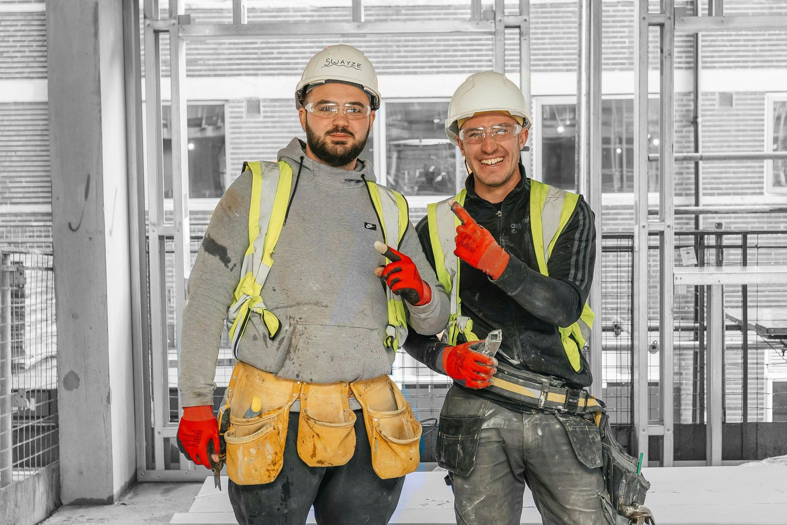 Two construction workers smiling and pointing at each other wearing full protective clothing