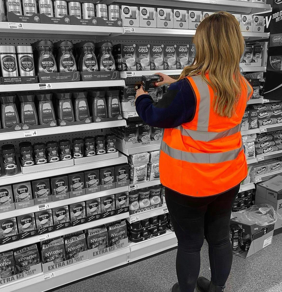 Retail worker with long hair and hi vis jackets replenishing a supermarket shelf